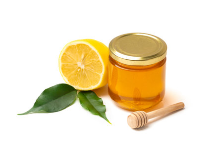 Honey in a glass jar and lemon with leaves on a white background. Closed jar with honey and honey stick on isolationの写真素材