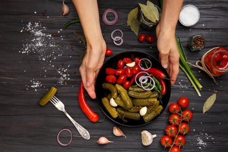 A plate with canned vegetables in hands on a dark background. Pickled cherry tomatoes and gherkins in a black plate top view. Pickled vegetable appetizer. home preservation. Preparations for the winter.の写真素材
