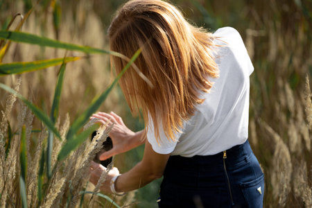 Girl in a white T-shirt in the park. The girl photographs spikelets in the field. hobby. Red-haired girl in a field of spikelets. A woman photographs nature.の写真素材