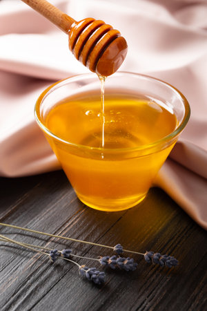 Honey is poured from a wooden dipper into a transparent glass bowl and lavender flowers on a dark wooden and neutral textile background. Conceptual composition of pouring honey.の写真素材