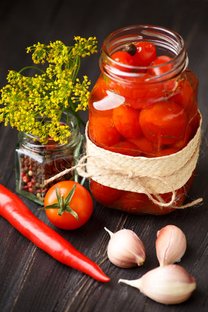 Pickled cherry tomatoes in an open glass jar, chili peppers, garlic, seasonings, spices and herbs for marinade close-up on a dark wooden background. home preservation.の写真素材