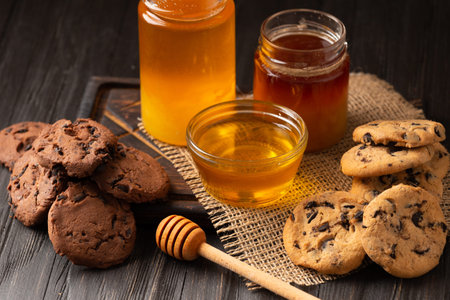 Honey in open jars and in a bowl, dipper and homemade cookies with chocolate chips on a wooden background. Homemade cakes and honey for breakfast. Composition of craft chocolate cookies and honey.の写真素材