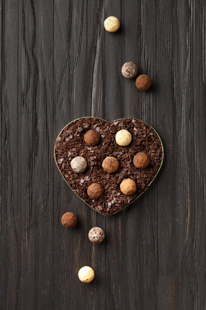 Chocolate truffles of different types in a heart-shaped box with chocolate chips on a dark wooden background, concept photo for Valentine's Day. Candies in chocolate chips on a dark background.の写真素材