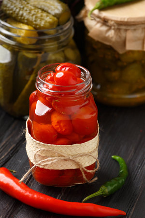 Canned cherry tomatoes and cucumbers in open jars, hot peppers on a dark wooden background. home preservation.の写真素材