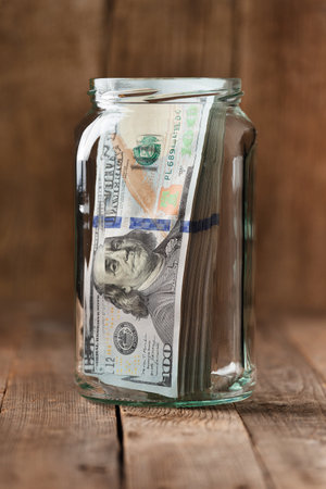 Dollar bills in a glass jar on a wooden background, a stack of 100 dollars banknotes in a jar. The concept of saving money, accumulating funds.の写真素材