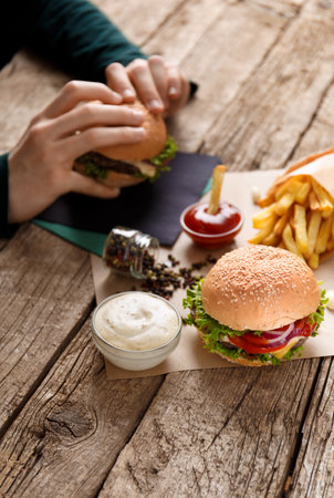 Appetizing hamburger with juicy cutlet, french fries, sauces on a rustic wooden background, burger in hands in the background. fast food concept.の写真素材