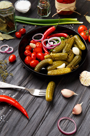 Homemade canned tomatoes, cucumbers, chili peppers in a round black plate, gherkin on a vintage fork on a dark wooden background.の写真素材
