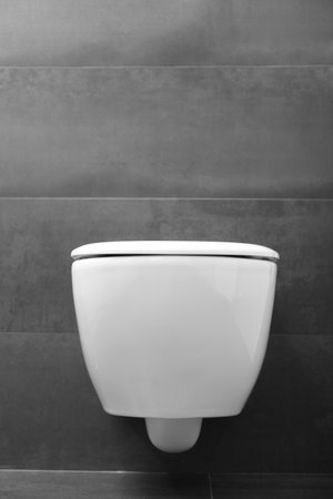 A modern wall-mounted white toilet with a closed lid and a shiny chrome flush button against a black wall. Toilet bowl in the interior of the bathroom.の写真素材