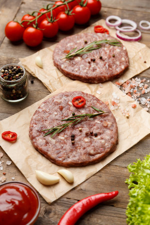 Raw burger patties with spices and rosemary on parchment, sauce, fresh vegetables, hamburger ingredients on a wooden background.の写真素材