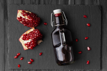 Freshly squeezed pomegranate juice in a glass transparent bottle, broken pomegranate and pomegranate seeds on a black flat stone plate on a dark wooden background top view.の写真素材