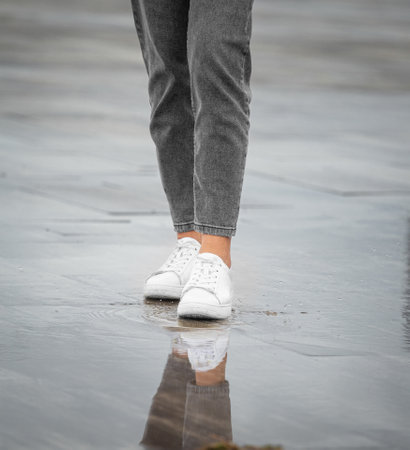 Legs of a girl in jeans and white sneakers walk in rainy weather on a street in the city, raindrops, water splashes, reflection in puddlesの写真素材