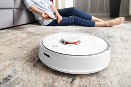 A girl sits on the floor in the living room and controls a robotic vacuum cleaner using a smartphone. smart home. The concept of smart home appliances, housework and technology.の写真素材