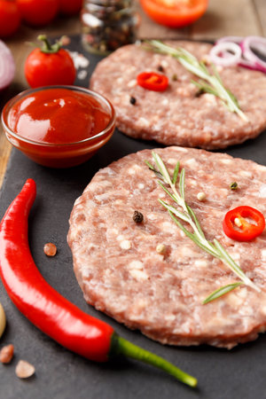 Raw fresh ground beef burger patties, rosemary, vegetables and spices on a black stone plate on a wooden background. Ingredients for a hamburger.の写真素材