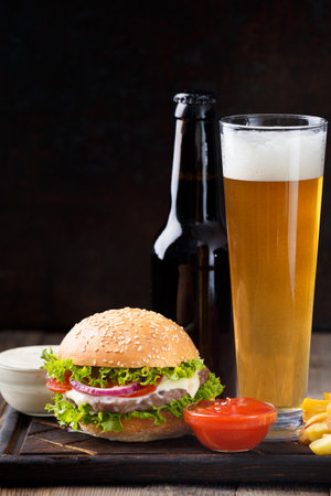 Burger, beer in a bottle and in a glass mug, French fries, sauce on a dark background, fast food concept.の写真素材