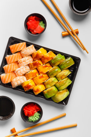 A set of bright multi-colored sushi rolls with shrimp, salmon, avocado in plastic packaging, Chinese chopsticks, sauce, ginger on a white background, top view. Sushi deliveryの写真素材