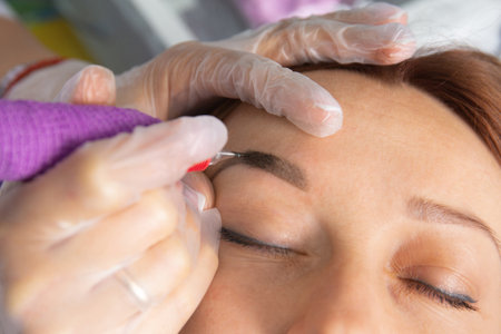 Beauty master performs permanent eyebrow makeup in a beauty salon, close-up. Hands of a cosmetologist doing microblading of eyebrows. Tattoo on the face.の写真素材