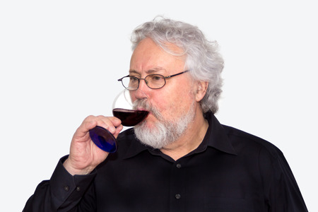 A gray haired bearded senior is tasting red wine isolated on whiteの写真素材
