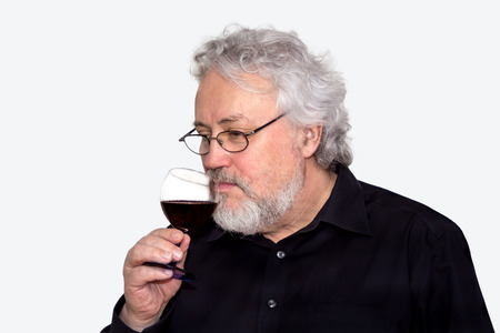 A gray haired bearded senior is tasting red wine isolated on whiteの写真素材