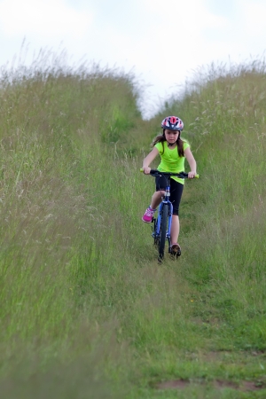 Girl teens in neon yellow shirt and a red helmet riding a bike down the meadow pathの写真素材