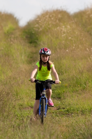 Girl teens in neon yellow shirt and a red helmet riding a bike down the meadow pathの写真素材