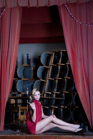 Actress young lady in red dress sits sad with a cigarette and a glass of alcohol on a dusty old stageの写真素材