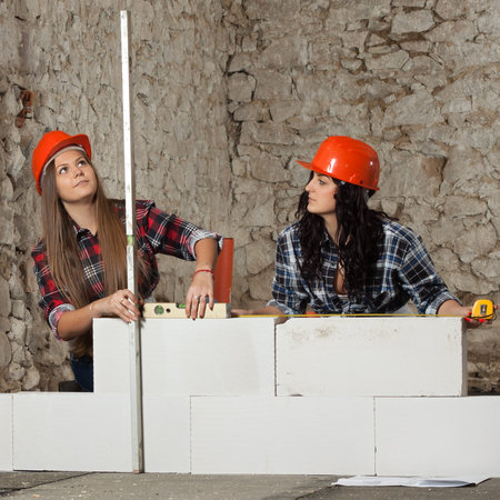 Two young long-haired woman repairing the old house and build a new wall from blocks in accordance...