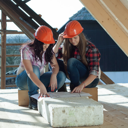 Two young women workers sitting on under construction roof, and control building in accordance with drawingの写真素材