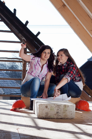 Two young women workers sitting on under construction roof, and control building in accordance with drawingの写真素材