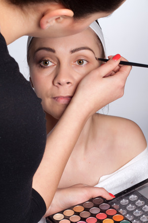 Beautician holding in one hand palette cosmetic shadows, and using a brush to apply color to the eyebrows of young brunette woman with headband on her headの写真素材