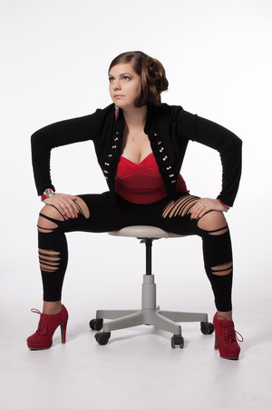 Young woman in red shirt, modern jacket, leggings with holes, red high heels, with exceptional haircut sitting on a swivel chair and posingの写真素材