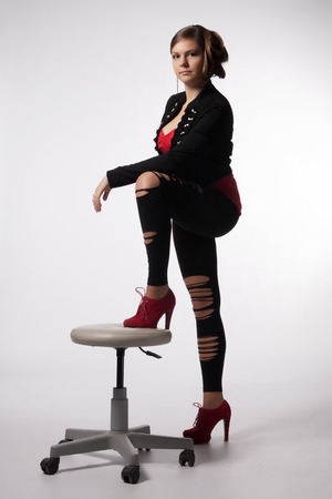 Young woman in red shirt, modern jacket, leggings with holes, red high heels, with exceptional hairstyle standing with your foot resting on a swivel chairの写真素材