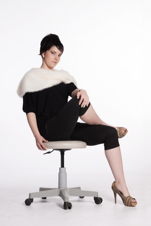 Portrait of a young woman in black with white fur collar sitting on a swivel chairの写真素材