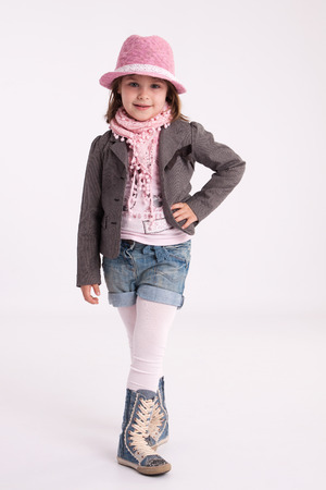Little girl preschooler model in pink hat, coat, shorts and sneakers high with a scarf on her neck posing in studioの写真素材