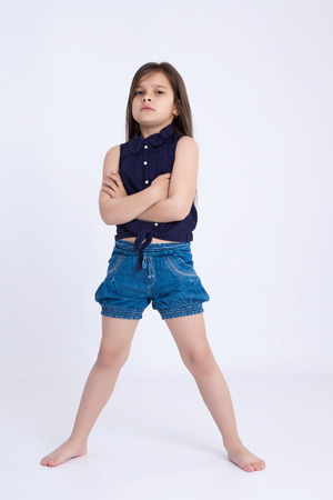 Little preschooler girl in denim shorts and a blue shirt standing with arms crossed and looking angrilyの写真素材