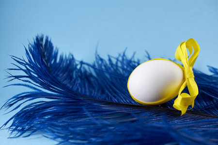 A white egg in a blue feather with yellow ribbon on a blue background. Spring concept.の写真素材