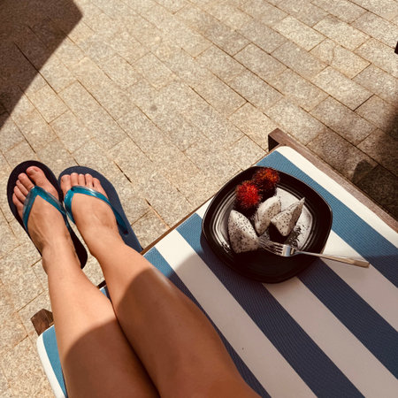 A close-up of the legs of a young woman in blue flip-flops lying on a sun lounger. A plate of exotic...