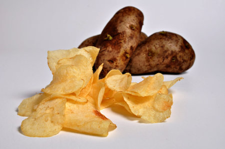 a pile of potato chips sit on a plain white background with a couple of potato's in the background.