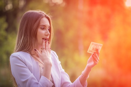 Excited curly young woman holding cash, in the parkの写真素材