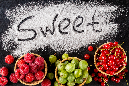 The word sweet is written on a dark background with the help of powdered sugar next to berries. raspberries, gooseberries, red currants. Top view, flat lay. Sweet, stylish desserts conceptの写真素材
