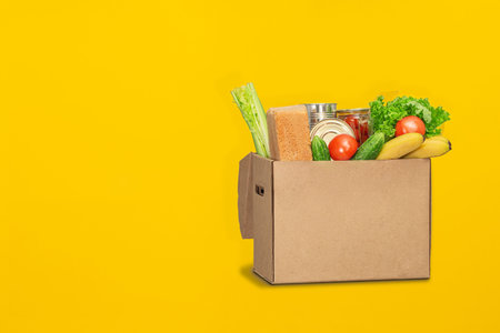 Donation box with food on a yellow background. Coronavirus food delivery. Volunteer brings a box of...