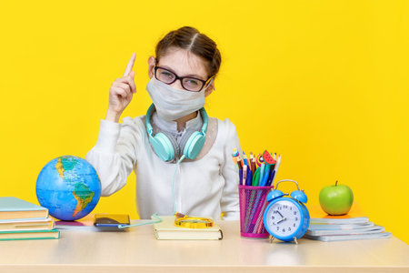 The schoolgirl at her Desk in a medical mask on yellow background