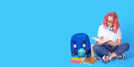Young female student reading books while sitting on floor with backpack over blue color background. female student sitting with books, headphones. Long wide banner. education concept, copyspaceの写真素材