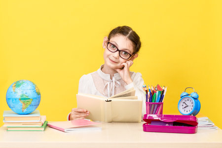 Back to school. The schoolgirl sits at the desk and thinks about the decision of the task on yellow background. Schoolchild in class. The new school year. Child education concept.の写真素材
