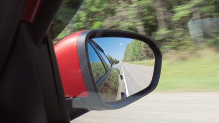 Driving a vehicle on highway at sunny day with view from side mirror. View in the rear view side mirror of a automobile, driving a red car along the road.の写真素材