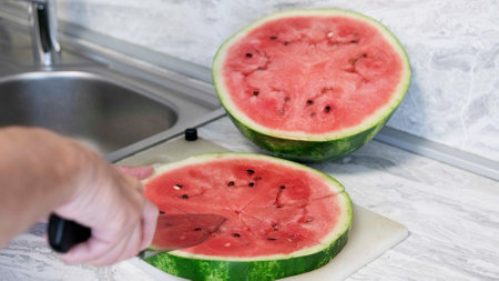 Man in home kitchen while cutting a ripe red watermelon in slices. The man use knife split the watermelon on on cutting board. Healthy food concept, season fruit, summer fruitの写真素材