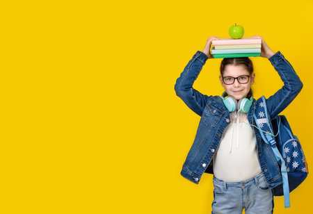 Back to school concept. Little happy girl in eyeglasses and headphones with stack of books on her head on yellow background. A schoolgirl holds a textbooks on her headの写真素材
