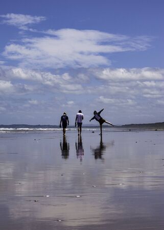 Two boys walking on a beach and a girl making a star pose with their reflections on the glossy sandの写真素材