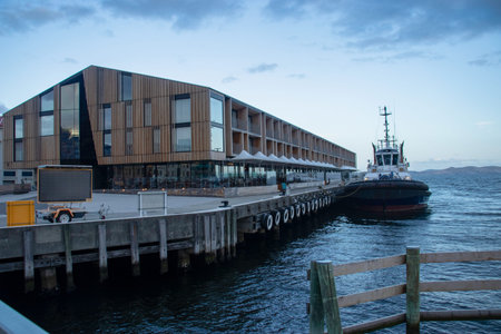 Large wooden building on a town pier with cloudy skies and a boat docked next to itのeditorial素材
