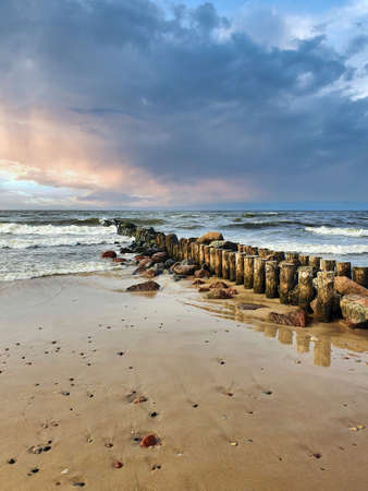 Empty sandy beach on the Baltic sea coast with calm waves rolling on old wooden breakwater under dramatic cloudy sky with stormy clouds at sunset. With no people peaceful seascape.の写真素材