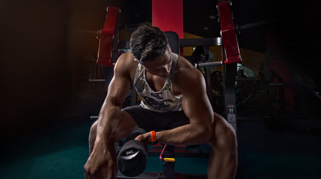 athlete strains biceps in the gym with dumbbells, creative photo, contours, muscles stand out with a watch on his arm, in an empty gym, beautiful body, physique, relief, sexy guyの写真素材